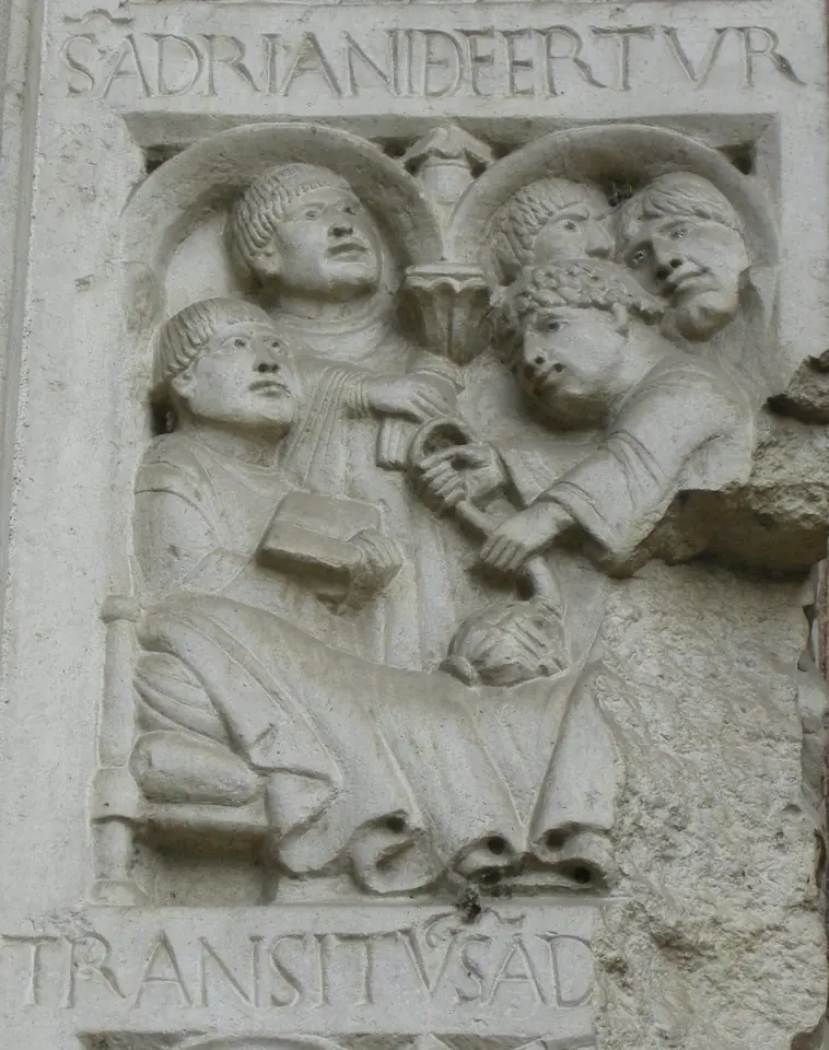 Romanesque stone relief of a dying man lying in bed while monks gather around him, one holding a book, with a Latin inscription TRANSITUS S. AD below