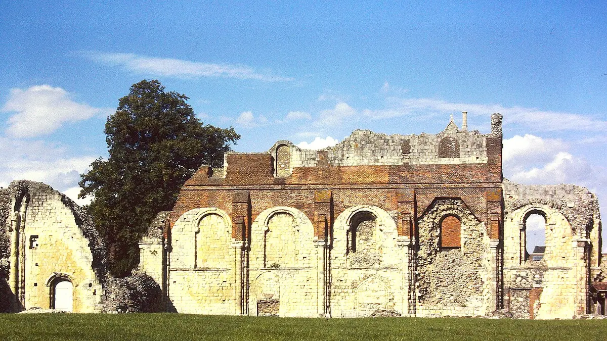 Golden stone and red brick walls of a ruined medieval abbey church with round arches, standing on green lawn under a blue sky