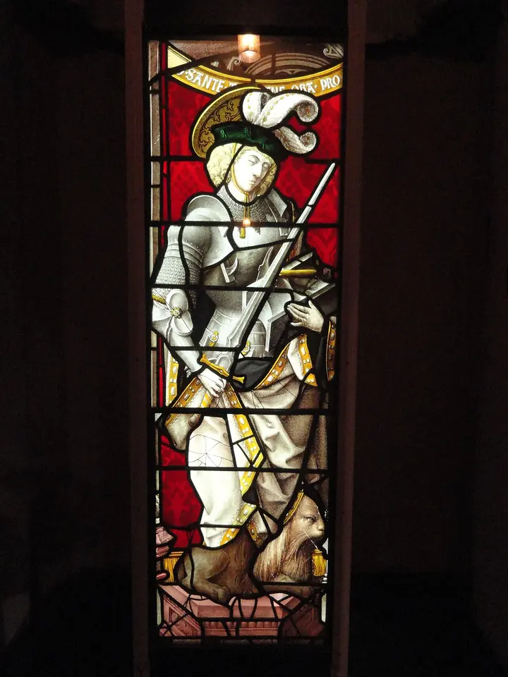 Stained-glass panel lit from behind, showing St. Adrian in white armor and a plumed hat with a sword, a lion crouched at his feet