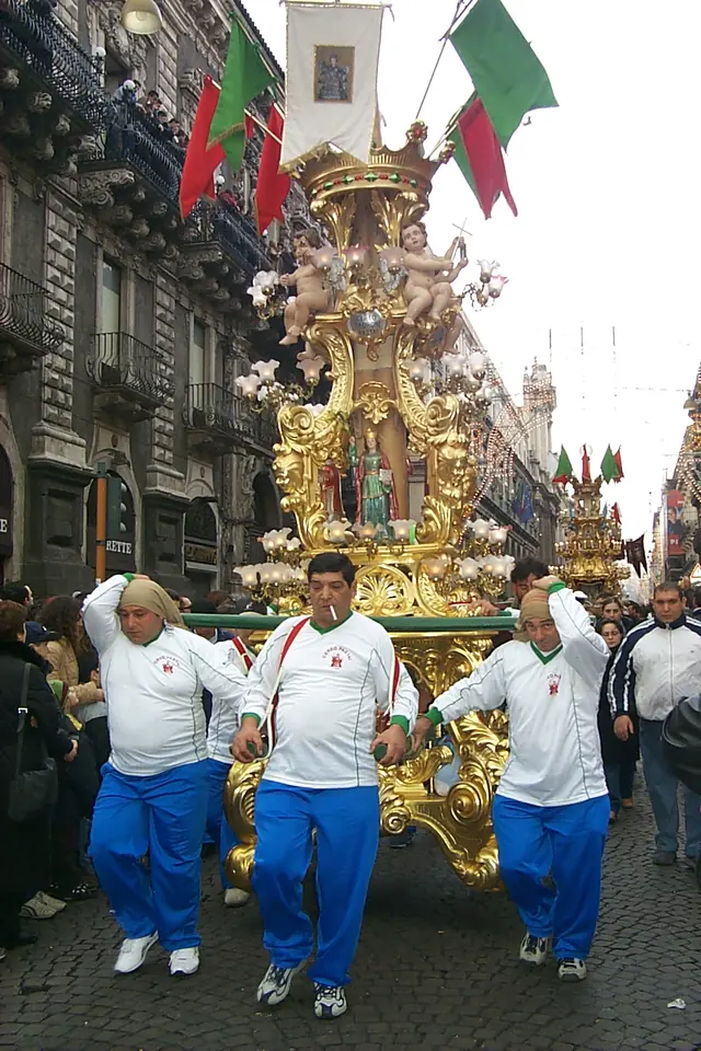 A tall gilded wooden candle-stand carved with cherubs and scenes, carried on the shoulders of men in white shirts and blue trousers down a city street hung with flags