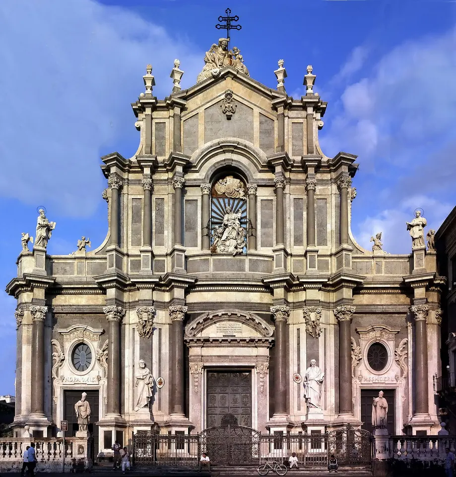 The tall Baroque stone facade of Catania Cathedral with columns, statues and a cross on top under a blue sky