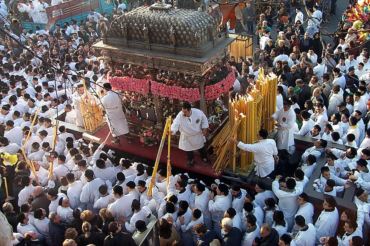 A silver processional carriage decorated with red flowers and tall candles moving through a dense crowd of devotees dressed in white