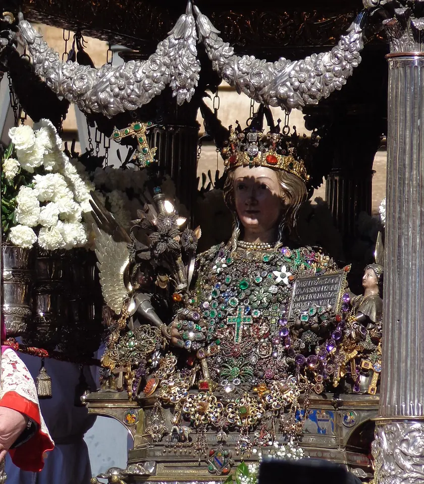 A silver reliquary bust of St. Agatha with a painted face, a jeweled crown and a chest covered in jewels, holding a small inscribed tablet, framed in silver