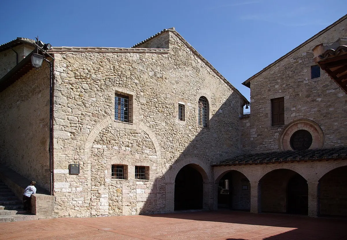 The plain stone church and convent of San Damiano with arched porch and a paved courtyard under a blue sky