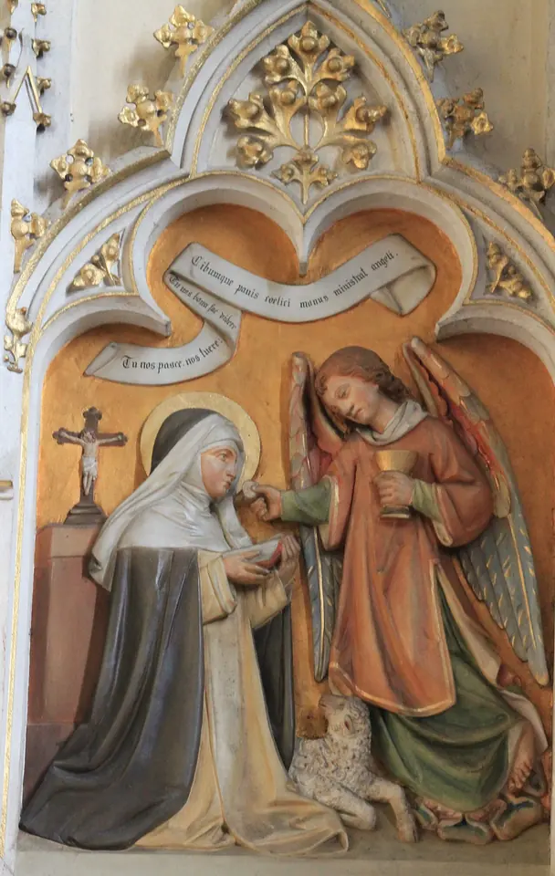 Painted Gothic Revival relief of a kneeling Dominican nun receiving the Host from a standing angel holding a chalice, with a crucifix on the wall and a lamb at her knees