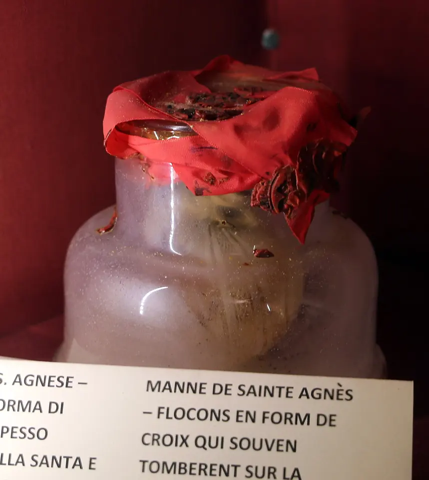 A small glass jar sealed with red cloth and wax, holding white grains, displayed behind glass with printed labels in Italian and French