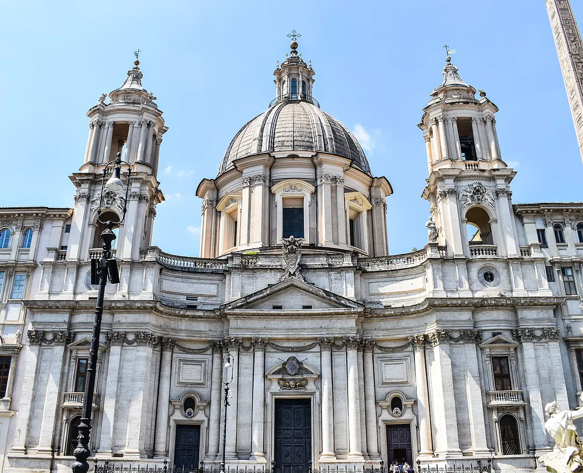Curving Baroque church front of pale stone with a central dome between two bell towers, under a blue sky