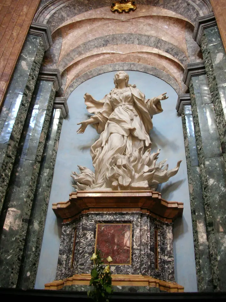 White marble statue of St. Agnes standing on carved flames with her arms open and her robe blowing, in a niche of green marble columns above an altar