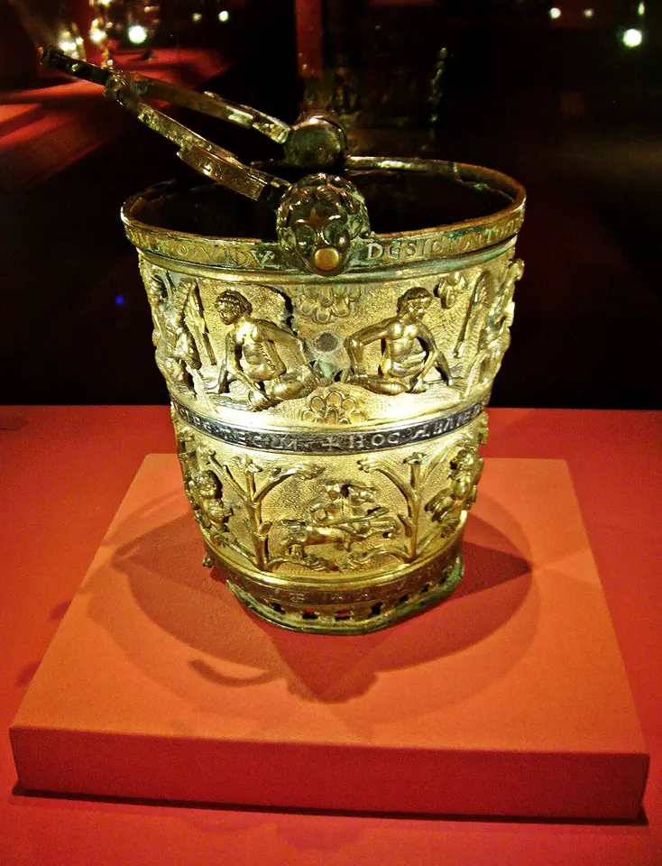 Gilded bronze holy water bucket with a handle, covered with relief figures in arches, displayed on a red stand in a dark museum case