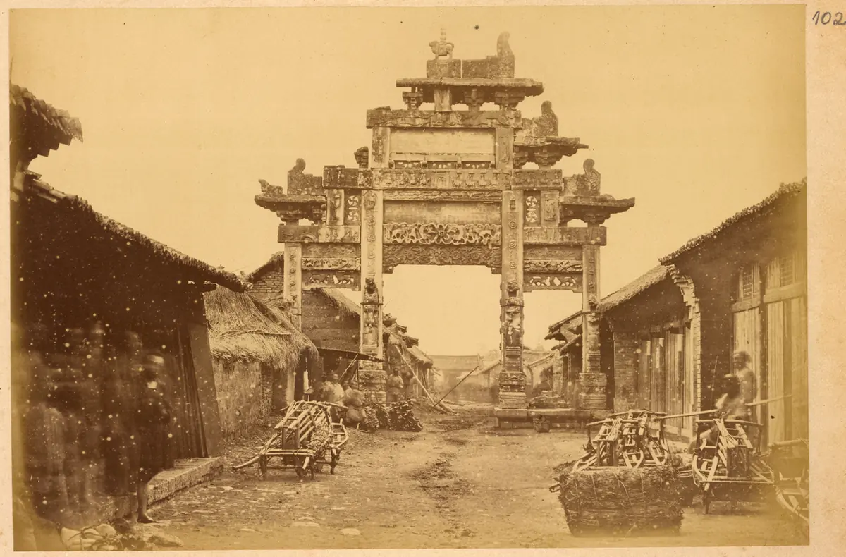 Old sepia photograph of a tall carved stone gateway spanning a dirt street lined with low wooden shops and stalls