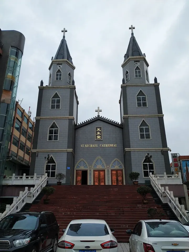 Gray twin-towered Gothic-style church with a red staircase leading up to its doors, among city buildings