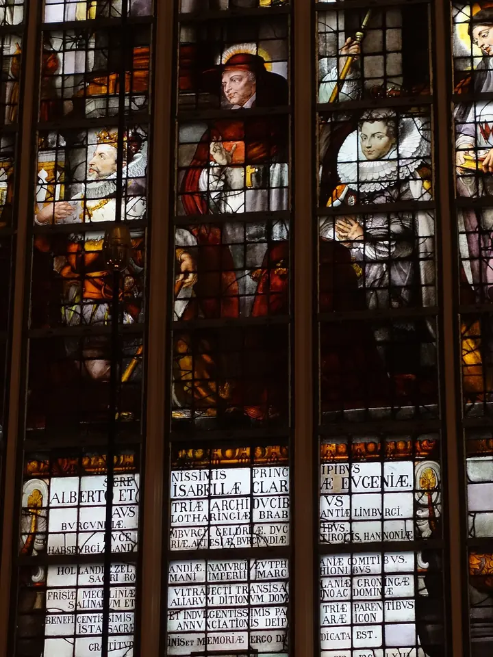 Detail of a tall stained-glass window showing a kneeling archduke, a saint in cardinal's red behind him, and a royal lady in a ruff, above panels of Latin inscriptions