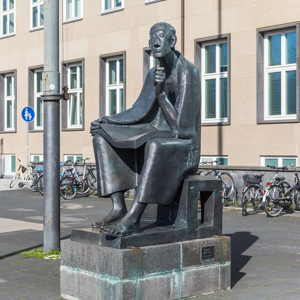 Bronze statue of a thin, seated friar with a long face resting his chin on one hand and an open book on his knees, in front of a university building