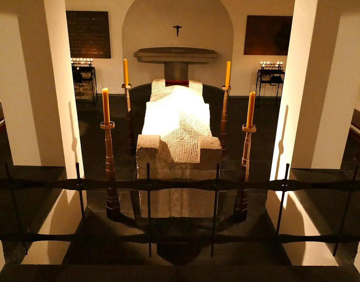 A pale stone Roman sarcophagus behind a low black railing in a vaulted crypt, with tall candles standing at its corners