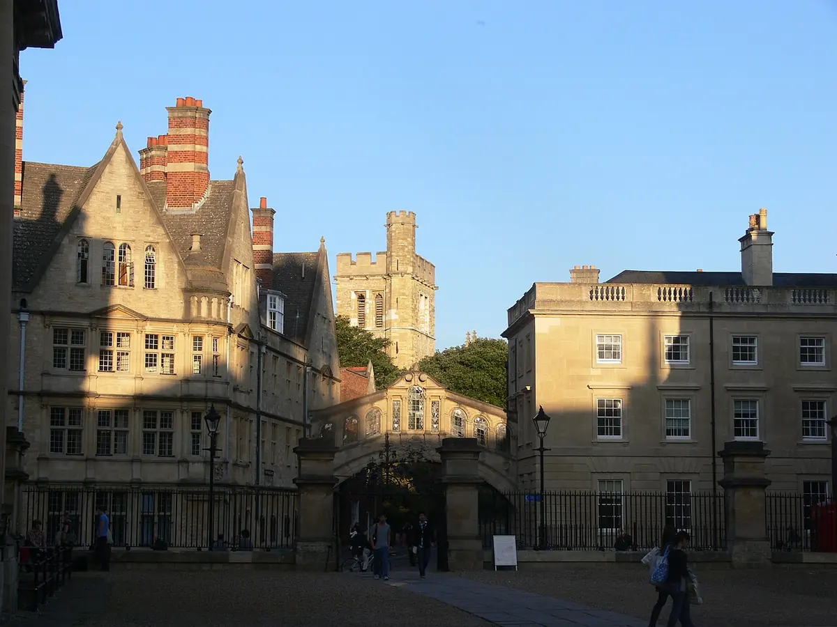Sunlit Oxford street with honey-colored college buildings, a covered bridge across the lane and a church tower beyond