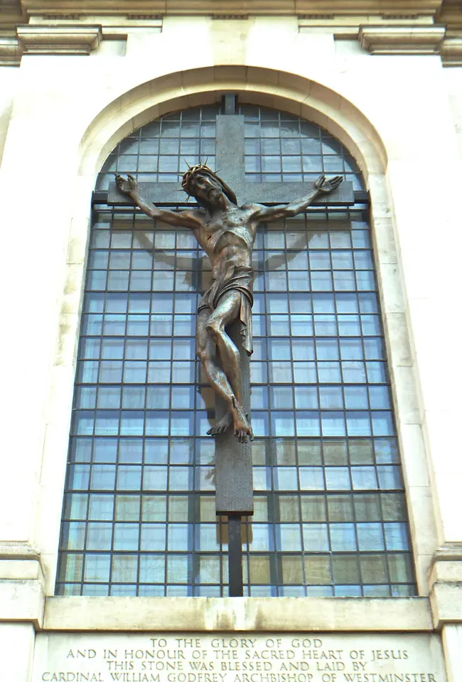 Bronze crucifix mounted on a tall arched window of a pale stone building, with an inscribed dedication stone below