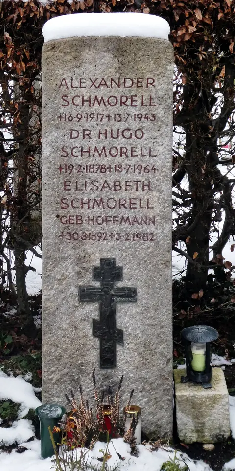 A tall pale stone grave marker carved with the names Alexander, Hugo and Elisabeth Schmorell above a Russian Orthodox cross, with snow and grave lights around it