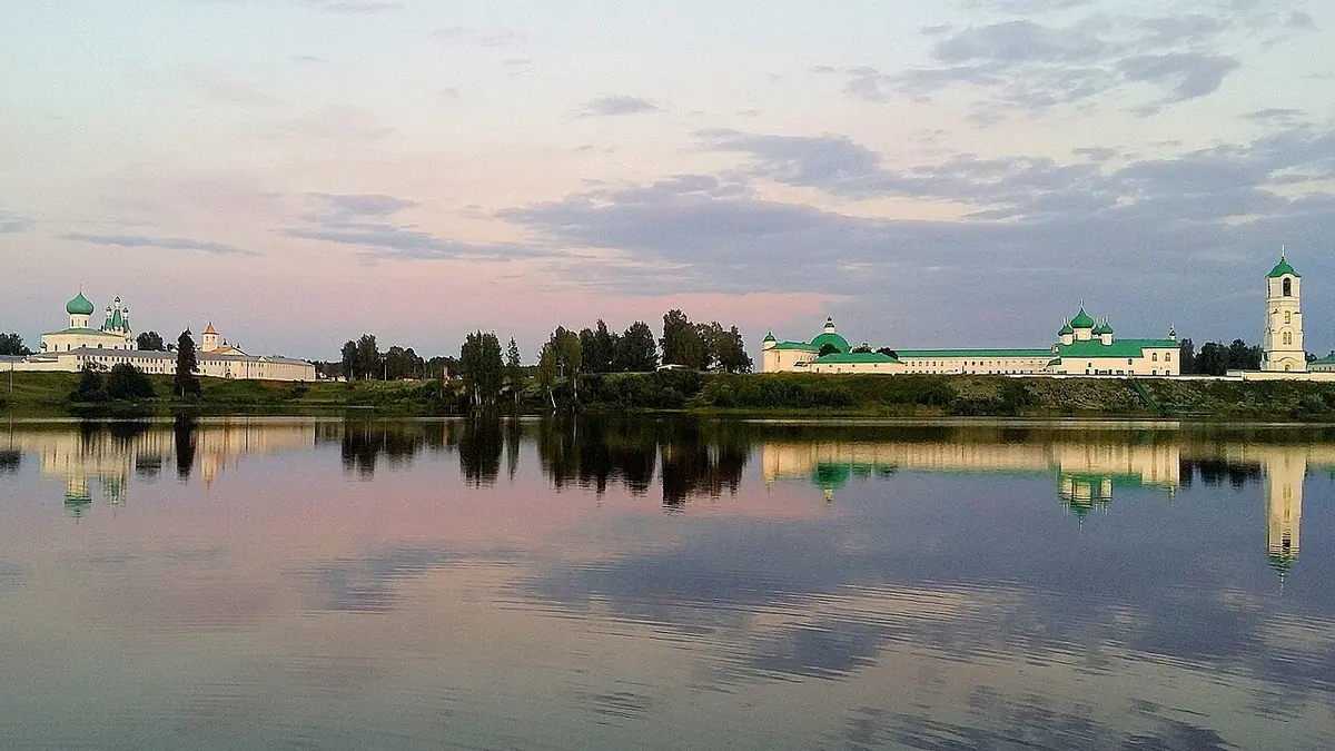 White monastery buildings with green roofs and domes reflected in a still lake under a pink evening sky