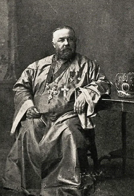 Old photograph of a seated, gray-bearded priest in a silk cassock with a pectoral cross and medals, one arm on a table beside a miter