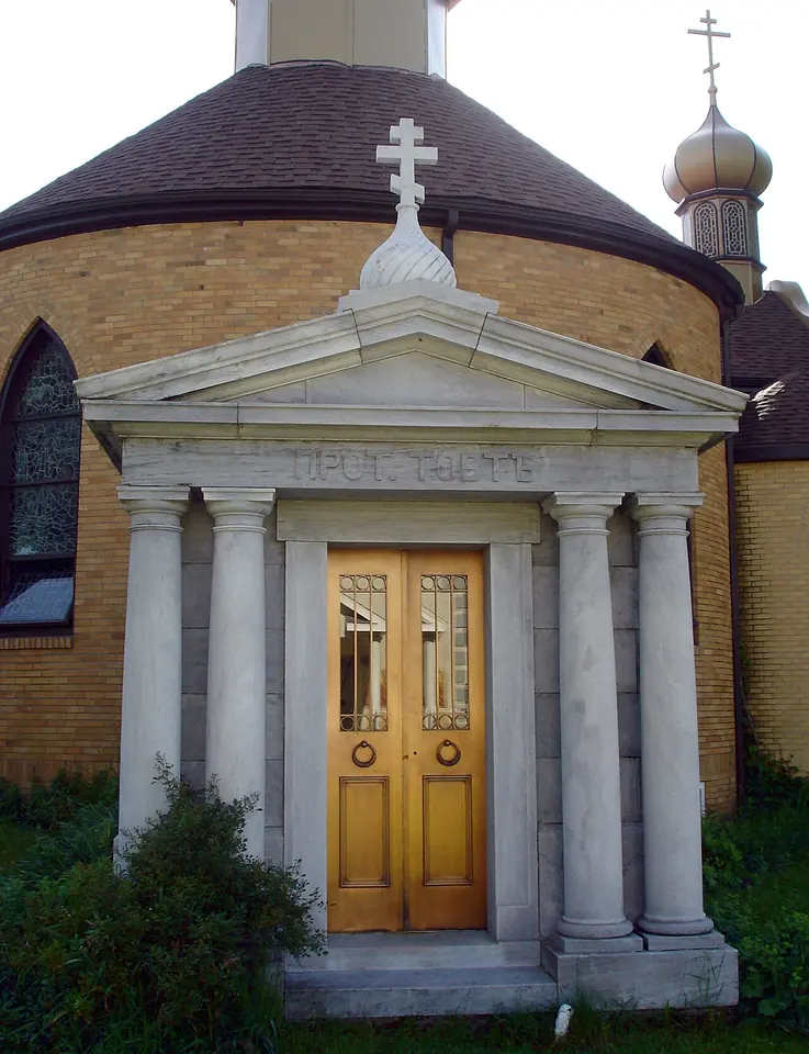 Small white classical mausoleum with columns and golden doors in front of a round brick church with a dome and onion cupolas