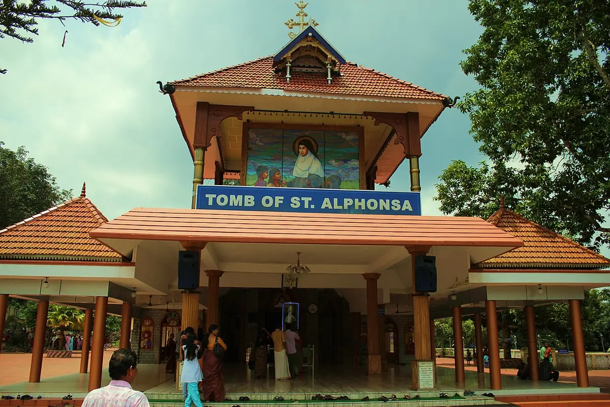 A tiled-roof chapel with a large portrait of St. Alphonsa above a blue sign reading "Tomb of St. Alphonsa," with pilgrims at the entrance