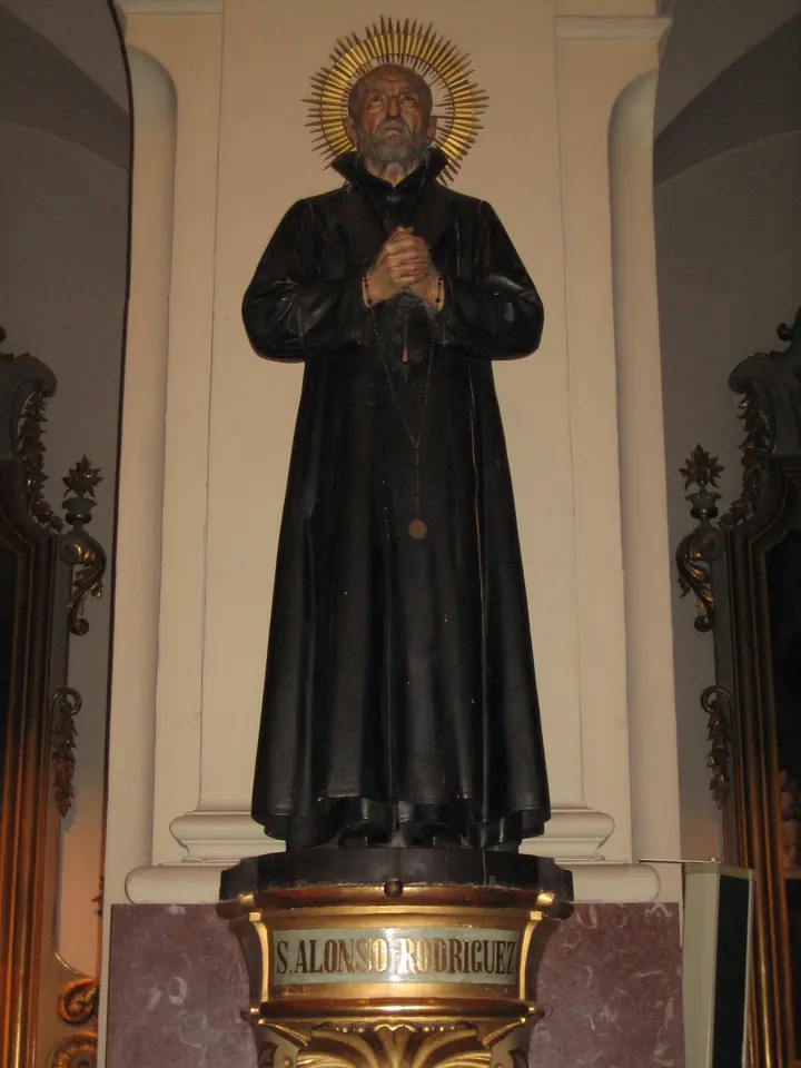 A dark carved wooden statue of St. Alphonsus Rodriguez with joined hands and a gilded halo, on a pedestal labeled S. Alonso Rodríguez