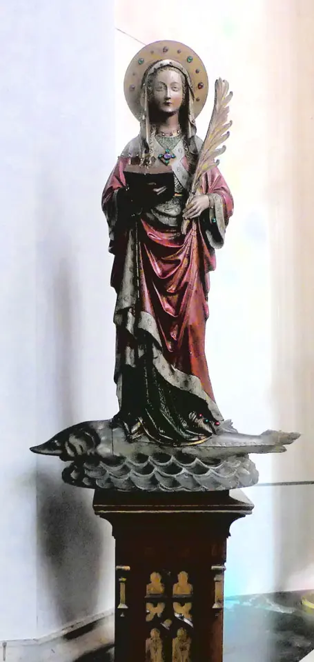 Painted wooden statue of St. Amelberga in a red cloak and white veil holding a palm, standing on a carved sturgeon on a tall pedestal