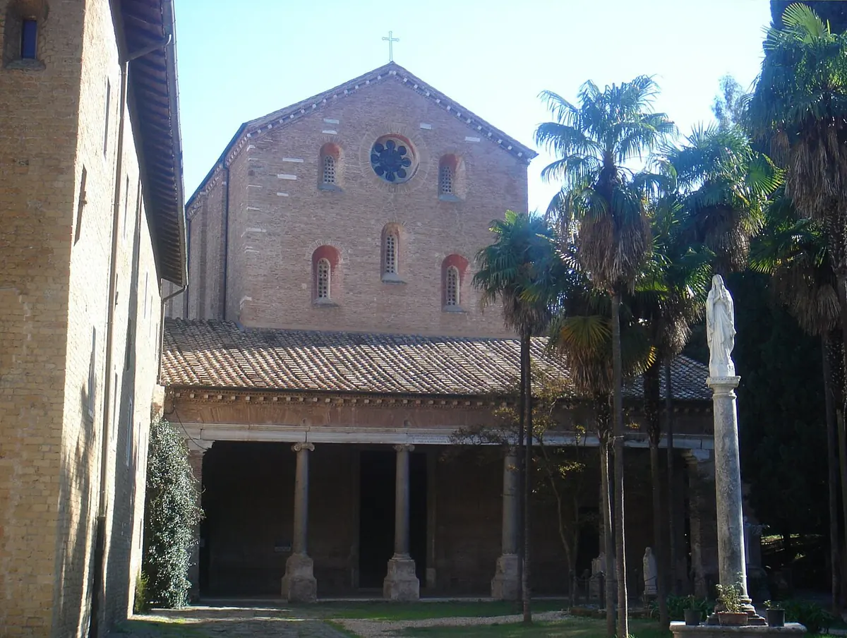 Plain brick facade of a Romanesque church with a round window above a colonnaded porch, palm trees and a white statue in the courtyard