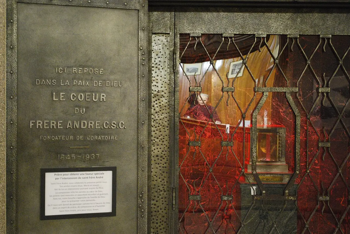 A bronze panel inscribed "Ici repose dans la paix de Dieu le coeur du Frère André C.S.C." beside a grille behind which a small lit reliquary glows red