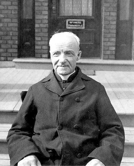 Black-and-white photograph of an elderly Brother André in a dark coat sitting on wooden steps in front of a brick building, smiling slightly