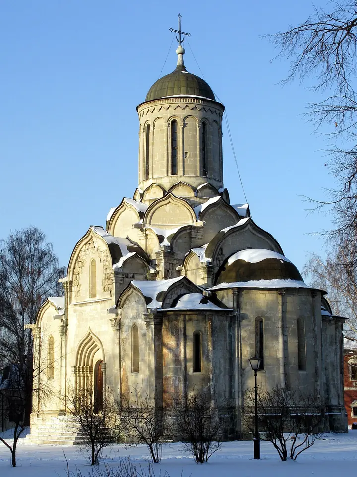 A small white stone church with stepped arches and a single helmet dome, standing in snow under a blue winter sky