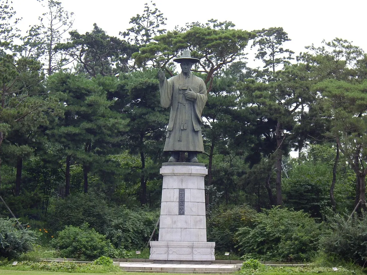 A tall dark statue of a man in a wide-brimmed hat and long robe on a white stone pedestal, among pine trees