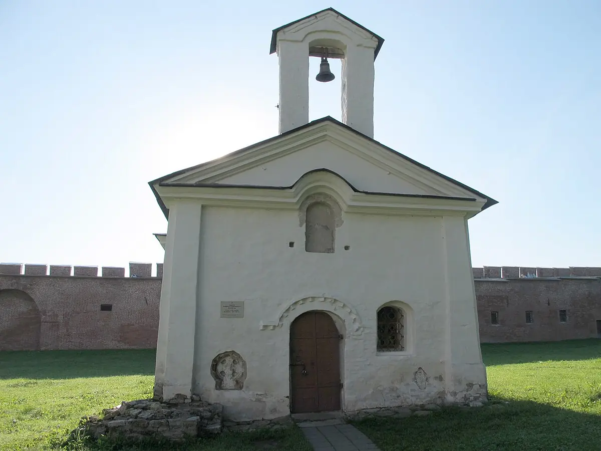 Small whitewashed stone church with a steep roof and a bell gable, standing on a lawn in front of the brick wall of the Novgorod Kremlin