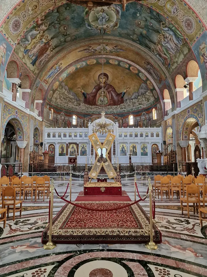 Interior of a large Byzantine-style church with painted domes, with a golden X-shaped reliquary cross standing on a red carpet before the iconostasis