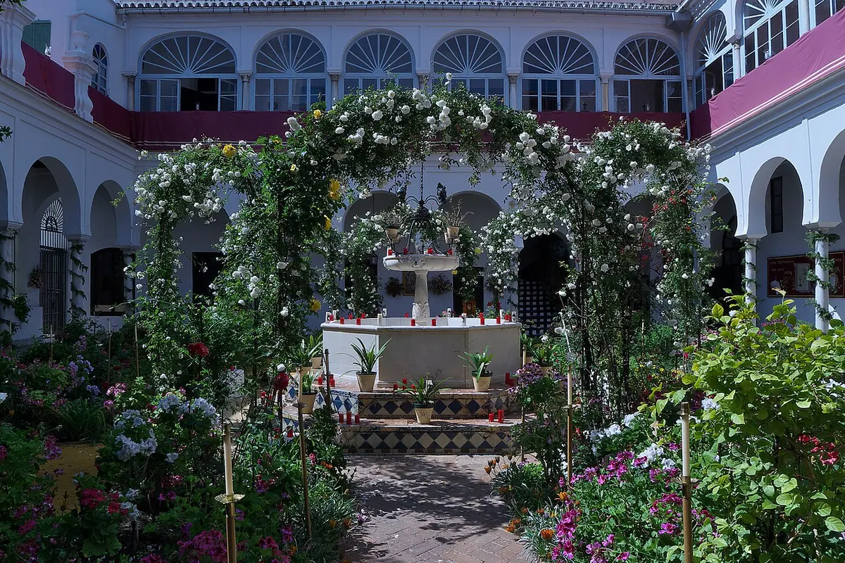 Sunny Andalusian courtyard with arched galleries, a central fountain and an arbor covered in white roses, full of potted flowers