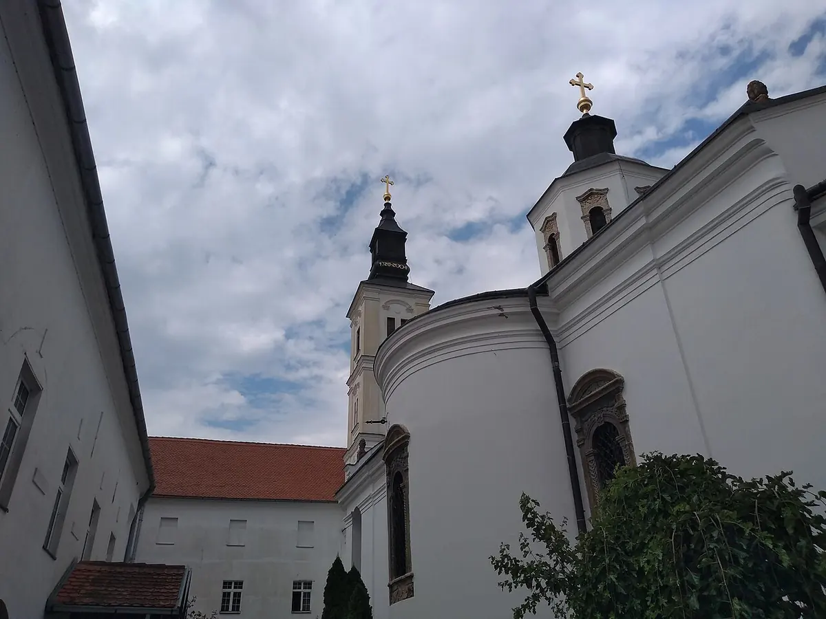 White monastery church with a Baroque bell tower and a gilded cross, seen from a courtyard with red-roofed monastery buildings