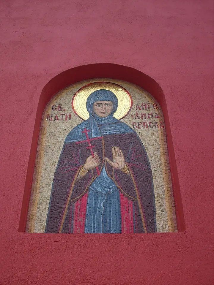 Gold mosaic in an arched niche in a red wall showing a nun in a blue veil and red mantle holding a small red cross and raising her right hand