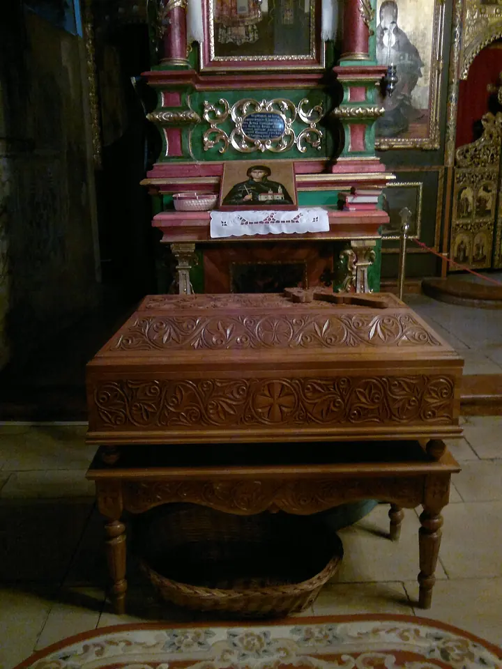 A carved wooden reliquary chest with a small wooden cross on its lid, set before an icon of a nun on a green and red painted stand in a dim church