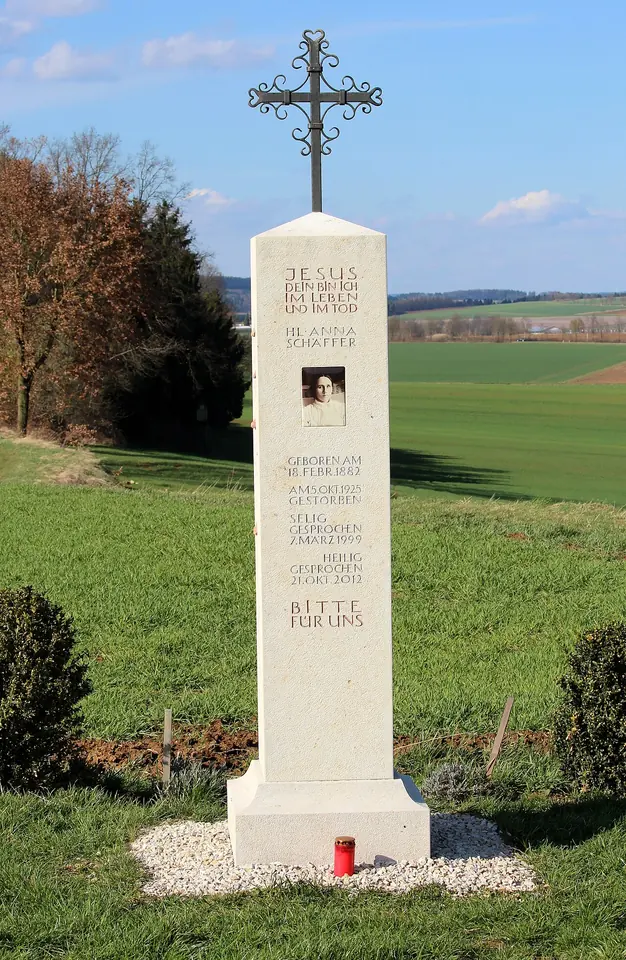 Tall white stone pillar topped with an iron cross, carved with lettering and a small photograph, standing at the edge of green fields