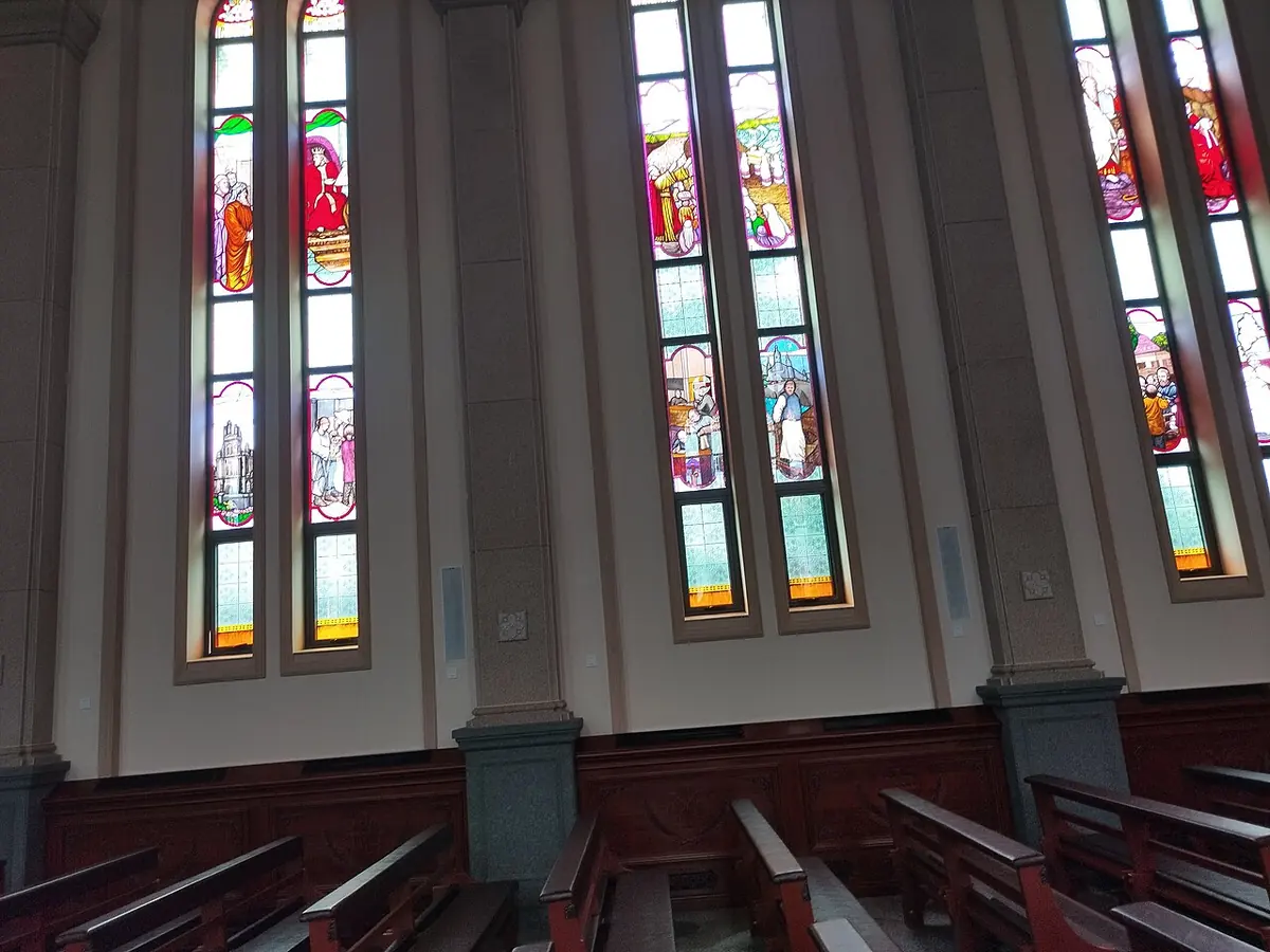 Tall narrow stained-glass windows with figures of saints in red, blue and gold along the nave of a church in northern China, above rows of wooden pews