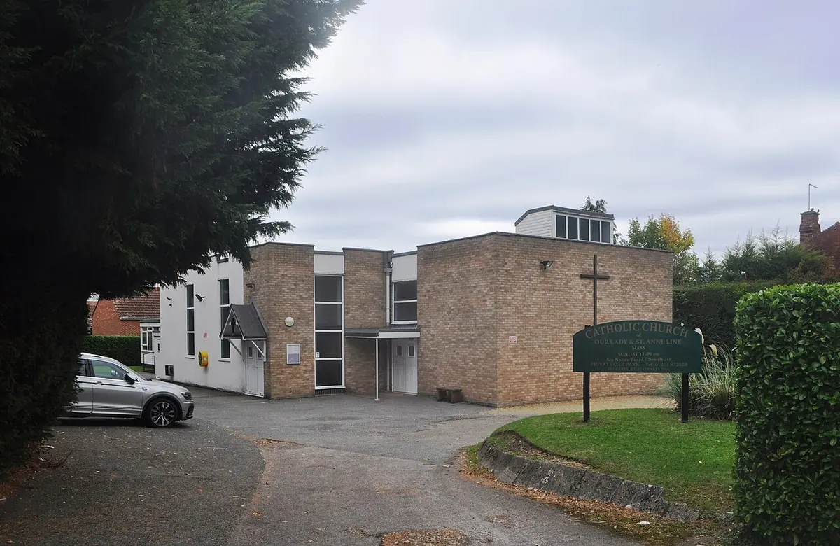 Modern brick church with flat roofs and a large cross on its side wall, seen across a parking area with a hedge and a sign board