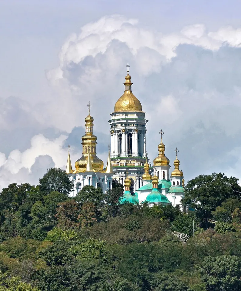 Golden domes and a tall white bell tower rising above green wooded hills over the Dnieper River in Kyiv