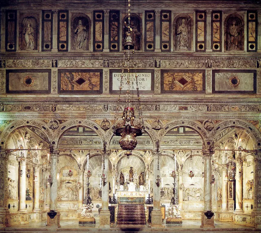 White marble Renaissance chapel with relief panels lining the walls and an altar tomb at the center under hanging silver lamps