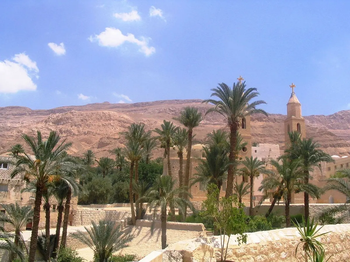 Palm trees and pale stone walls of a desert monastery with a bell tower, beneath bare reddish mountains