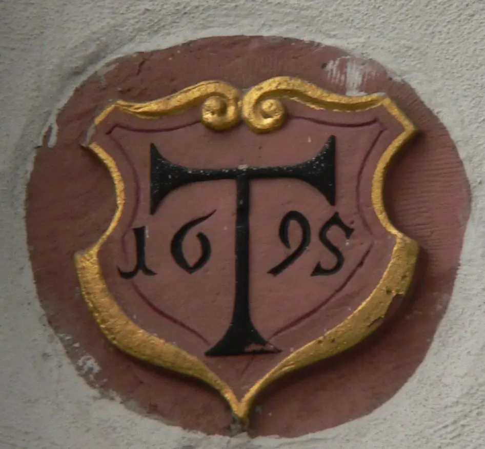 Carved shield on a red wall bearing a large black T-shaped tau cross in a gold border