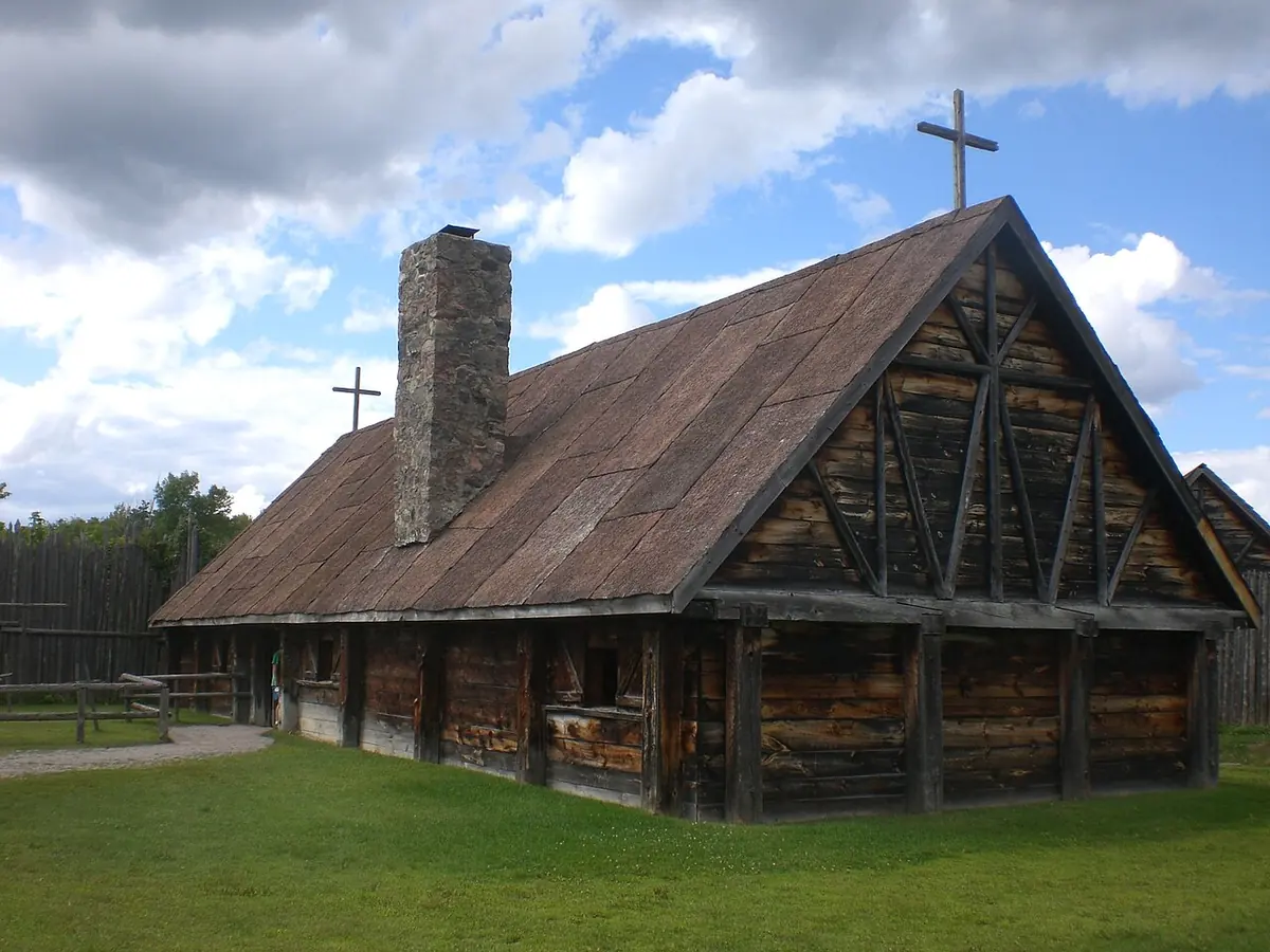 Long wooden chapel with a dark shingled roof, a stone chimney and a cross on the gable, on a green lawn