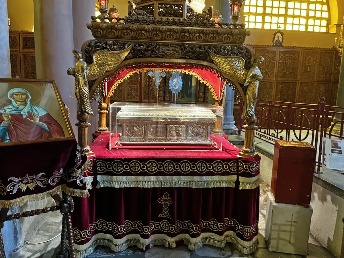 Silver and glass reliquary shrine under a carved wooden canopy on a table draped in red velvet, with an icon of the saint beside it, inside a church