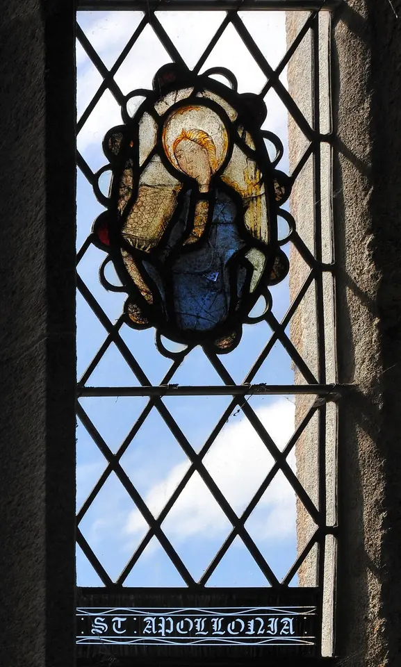A small oval panel of old stained glass showing a haloed woman in blue, set in a clear leaded window, with the words St. Apollonia painted below