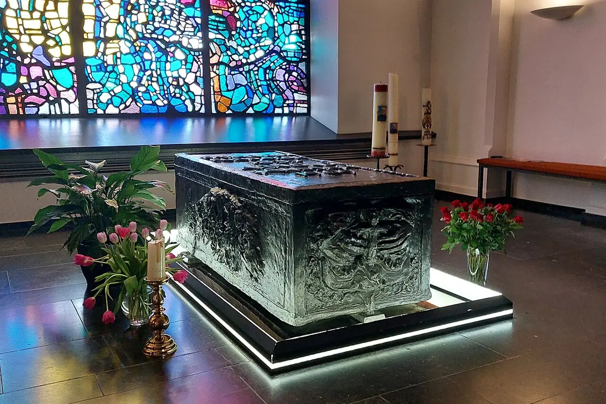 A dark metal sarcophagus decorated in relief on a lit platform, with flowers and candles, in front of a wall of brightly colored stained glass