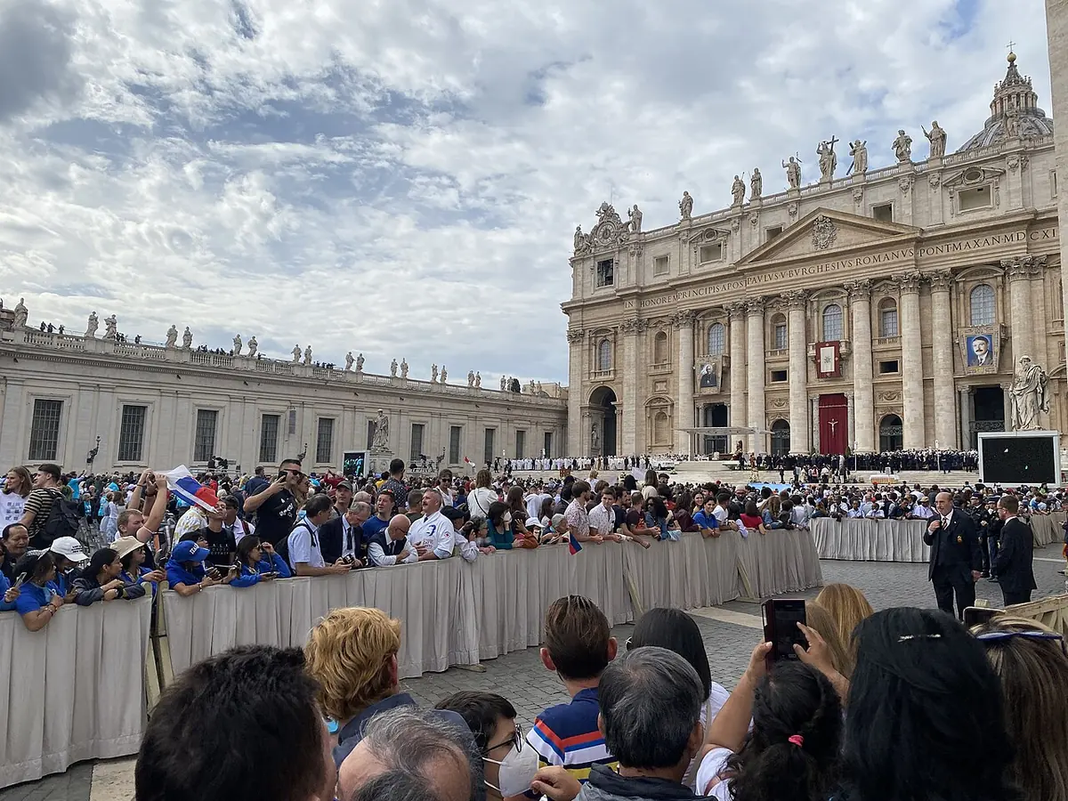 Crowds of pilgrims behind barriers in St. Peter's Square under a partly cloudy sky, with the facade of St. Peter's Basilica and its hanging tapestries in the background